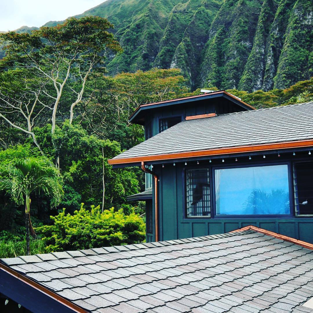 House with shingle roof surrounded by lush greenery and mountains.