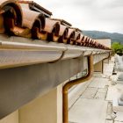 Clay tile roof with gutter and downspouts, set against a cloudy sky and distant hills.